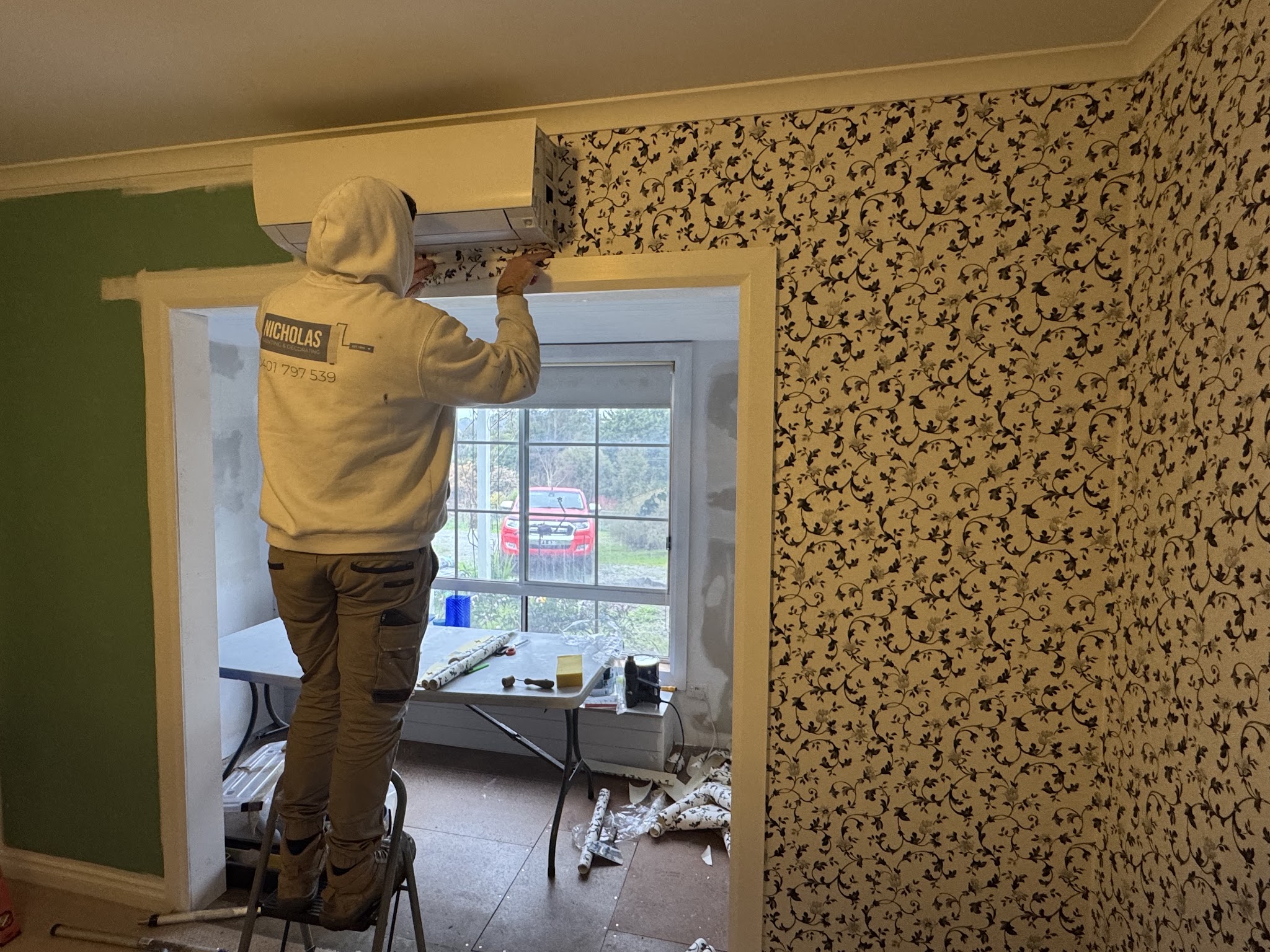 Bayley in a Nicholas Painting and Decorating hoodie on a step ladder, stripping old wallpaper from a wall